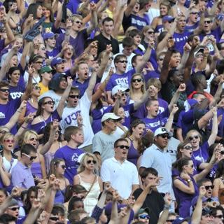 Dozens of cheering TCU fans raise their arm in a two-fingered "Go Frogs" hand sign at a crowded football game.